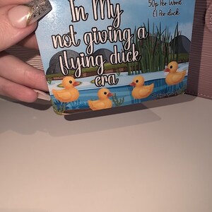 May include: A rectangular card featuring the phrase "In My not giving a flying duck era" and four cartoon ducks on a pond. The card also displays "50p Per Word £1 Per duck". The card is held by a person.