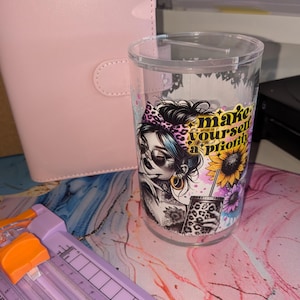 May include: Clear plastic tumbler featuring a graphic of a woman with sunglasses, a leopard print bow, and tattoos. The tumbler has the text "make yourself a priority" and a sunflower design. A pink planner and a purple paper cutter are in the background.