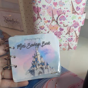 May include: A small, ring-bound "Mini Savings Book" with a castle illustration on the cover. The cover has the text "Mini Savings Book" and the Instagram handle "@PrettyDesignsByCo". A planner with a heart and Eiffel Tower pattern is in the background.