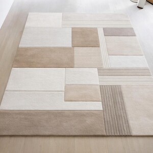 Puede incluir: Una alfombra rectangular con un diseño geométrico. La alfombra presenta un mosaico de formas rectangulares y cuadradas en tonos beige, crema y marrón. Algunas secciones tienen una textura acanalada, agregando interés visual al revestimiento del suelo.
