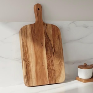 Może przedstawiać: Prostokątna drewniana deska do krojenia z uchwytem. Deska wykonana jest z jasnobrązowego drewna z widocznymi wzorami słojów. Uchwyt ma otwór do zawieszania. Deska opiera się o białą marmurową powierzchnię.