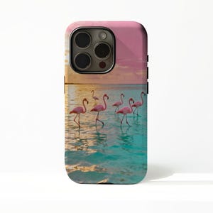 May include: A phone case depicting a tropical scene with pink flamingos in turquoise water under a pink and orange sunset. The case has a black camera lens and black side buttons.