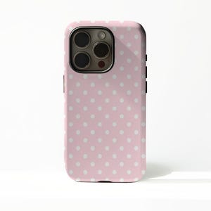 Pode incluir: Uma capa de telefone rosa com um padrão de bolinhas brancas. A capa tem um recorte para a câmera e botões laterais pretos. A capa é projetada para um smartphone.