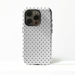 Könnte beinhalten: Eine weiße Handyhülle mit einem Muster aus gleichmäßig verteilten schwarzen Punkten. Die Hülle hat einen Kameraausschnitt und schwarze Seitentasten. Die Handyhülle soll das Telefon vor Kratzern und Beschädigungen schützen.