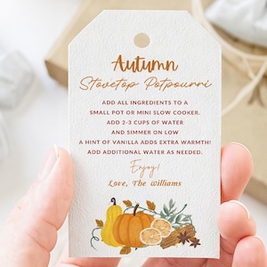 Op de afbeelding: Een wit label met de tekst "Autumn Stovetop Potpourri" en instructies voor het maken van potpourri. Het label bevat illustraties van pompoenen, sinaasappels, kaneelstokjes en steranijs. Het label wordt door een persoon vastgehouden.