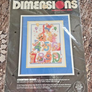 Może przedstawiać: Zestaw do haftu krzyżykowego Dimensions z wzorem "Counting Bears". Obraz przedstawia misie trzymające cyfry 1-9, z balonami. Zestaw zawiera instrukcje i materiały do ukończonego elementu o wymiarach 28x35cm lub 38x45cm.