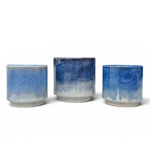 May include: Three cylindrical ceramic cups featuring a reactive glaze that transitions from a deep cobalt blue at the top to a lighter, muted blue and off-white base. Each cup has a distinct stepped design near the bottom, creating a clear rim.