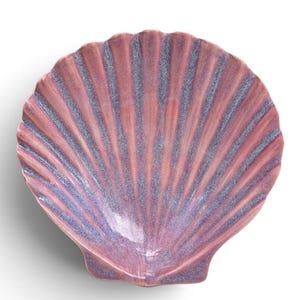 May include: A ceramic dish shaped like a scallop shell with a glossy finish. The surface features a gradient of pink and lavender hues, with darker purple tones accentuating the deep, radiating ridges that extend from the base to the scalloped edge.