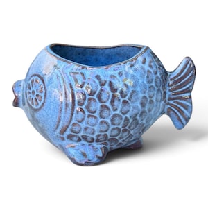 May include: A blue ceramic planter shaped like a fish with a textured scale pattern and dark brown accents along the fins, tail, and eye. The vessel features an open top and a wide, rounded body with a small mouth and a distinct tail fin design.