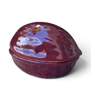May include: A glossy, deep plum-colored ceramic lidded container shaped like a walnut. The object features a smooth, reflective surface with organic, textured contours mimicking the natural shell of a nut, consisting of two distinct halves.