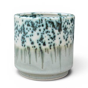 May include: A cylindrical ceramic cup featuring a white upper section with dark teal and black speckled patterns. The lower portion of the cup displays a smooth, light sage green glaze that drips downward, creating a layered effect on the exterior.
