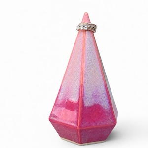 May include: A pink and purple ceramic ring holder in a geometric pyramid shape. The holder has a glossy finish and a ring with three clear stones is on top. The ring holder is a decorative item.