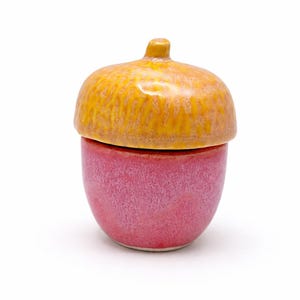 May include: A ceramic acorn-shaped container with a lid. The lid is a textured yellow-orange, and the base is a speckled pink. The container is designed to resemble an acorn, with a small stem on top.