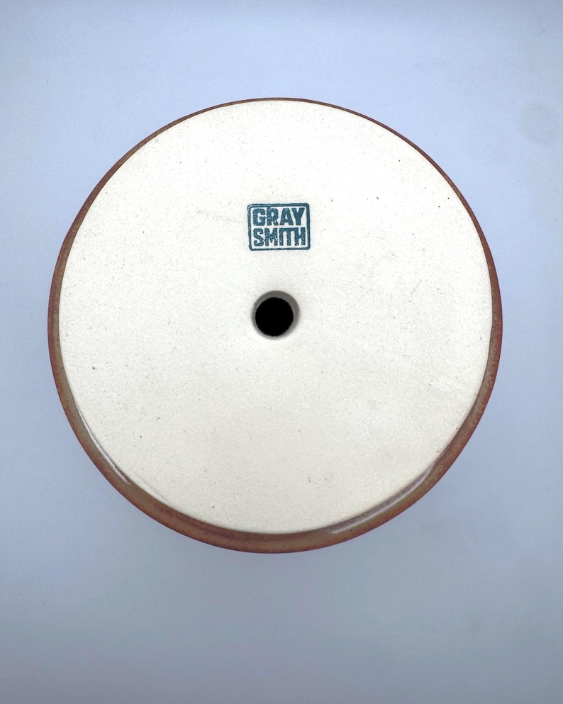 Puede incluir: Una pieza de cer&aacute;mica redonda, blanquecina, con un borde marr&oacute;n oscuro y un agujero central. El texto "GRAY SMITH" est&aacute; estampado en un cuadrado turquesa. Probablemente sea una base o tapa de cer&aacute;mica.