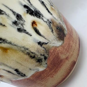 Puede incluir: Primer plano de un jarr&oacute;n de cer&aacute;mica con un dise&ntilde;o veteado &uacute;nico. El jarr&oacute;n presenta una base de color crema con vetas negras, marrones y beige. La parte inferior del jarr&oacute;n tiene una banda marr&oacute;n rojiza. El dise&ntilde;o le da al jarr&oacute;n un aspecto natural y org&aacute;nico.