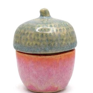 May include: A ceramic acorn-shaped container with a lid. The lid is a mottled green and blue, while the base is a gradient of pink and orange. The container is likely handmade and suitable for small items.