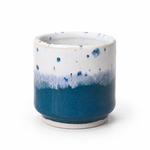 May include: A cylindrical ceramic cup featuring a two-tone glaze. The upper portion is white with scattered blue speckles, transitioning into a solid deep blue base. The bottom rim is unglazed, revealing a light beige, textured clay surface.