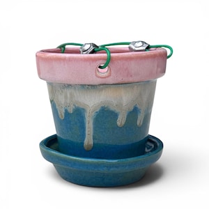 May include: A ceramic hanging planter with a pink rim and a blue body featuring a white drip glaze effect. The pot includes a matching blue saucer base and a green cord threaded through two holes in the rim for suspension purposes.