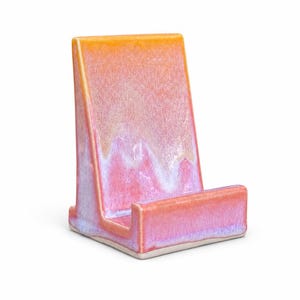 May include: A ceramic phone stand with a gradient of colors, transitioning from orange at the top to pink and purple at the base. The stand has a rectangular shape with a slot to hold a phone.