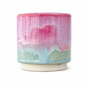 May include: A cylindrical ceramic planter featuring a reactive glaze with a vibrant magenta top that transitions into a soft blue and muted green base. The vessel sits on a contrasting, unglazed, off-white circular pedestal base with a speckled texture.