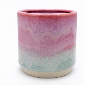 May include: A cylindrical ceramic mug with a gradient of colors. The top rim is a deep red, transitioning to pink, then lavender, and finally a light teal at the base. The base is a natural, unglazed beige.
