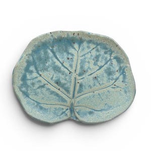 May include: A ceramic dish shaped like a rounded leaf with an irregular, wavy edge. The surface features a mottled teal and light blue glaze with a prominent embossed vein pattern running through the center and small dark brown speckles throughout.