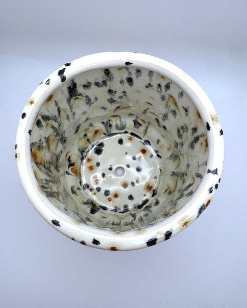 Puede incluir: Maceta de cer&aacute;mica con dise&ntilde;o moteado. El interior y el exterior presentan una base blanca con manchas negras y marrones. La maceta tiene forma redonda con un orificio de drenaje en la base.