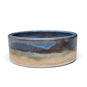 May include: A round ceramic bowl featuring a reactive glaze with a gradient of deep blue at the rim, transitioning into dark brown and earthy tan tones toward the base. The interior of the bowl is finished in a smooth, glossy blue glaze.
