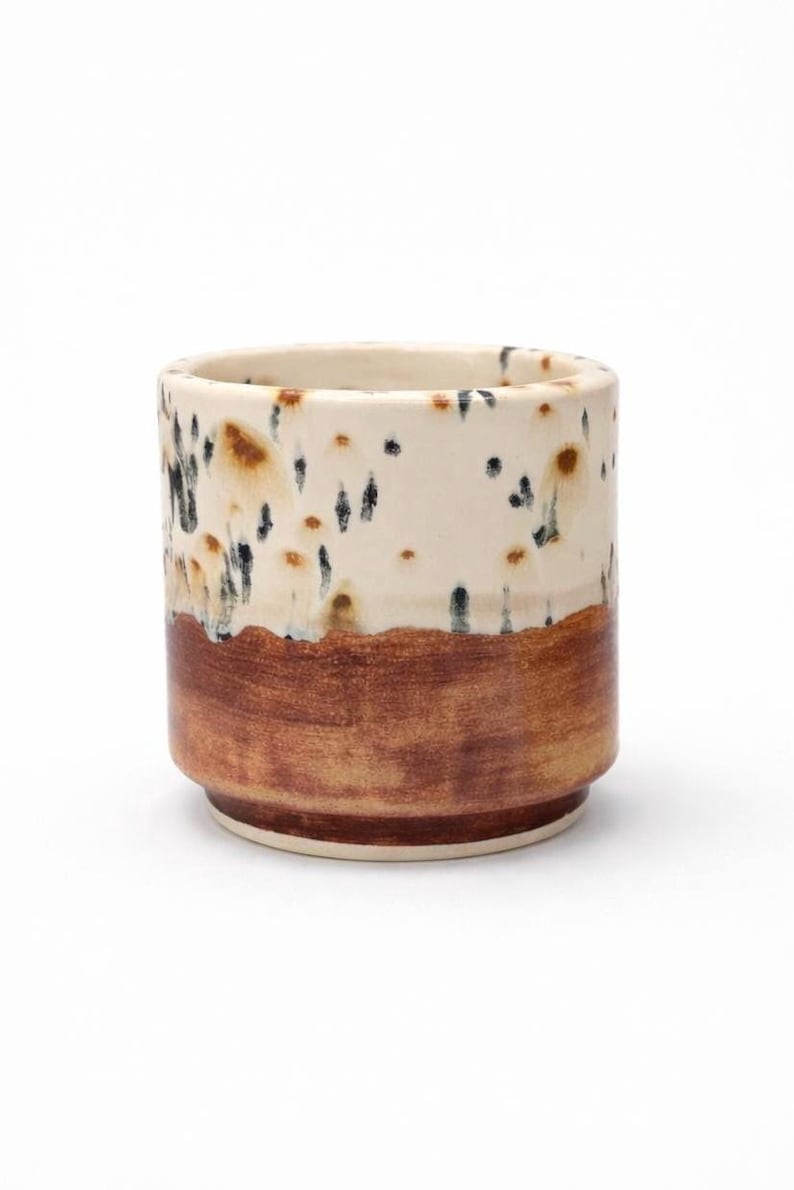 Puede incluir: Un vaso de cer&aacute;mica de forma cil&iacute;ndrica. La mitad superior es blanquecina con motas marrones y negras, mientras que la mitad inferior es de color marr&oacute;n oscuro liso. La base es de color marr&oacute;n claro.