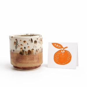 Puede incluir: Un vaso de cer&aacute;mica con un esmalte blanco y marr&oacute;n moteado, y una banda inferior marr&oacute;n. Junto a &eacute;l, una peque&ntilde;a tarjeta con una ilustraci&oacute;n naranja de un c&iacute;trico. El texto "Add a gift message card from my shop" est&aacute; en la parte inferior.