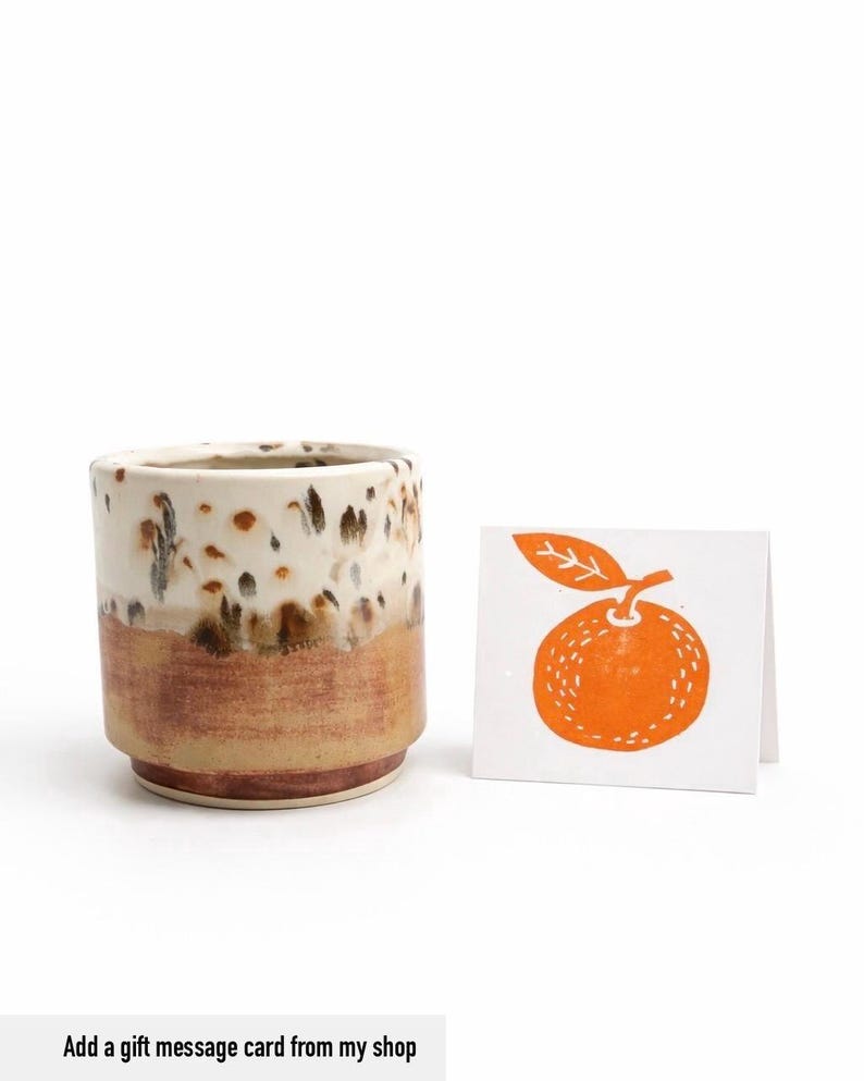 Puede incluir: Un vaso de cer&aacute;mica con un esmalte blanco y marr&oacute;n moteado, y una banda inferior marr&oacute;n. Junto a &eacute;l, una peque&ntilde;a tarjeta con una ilustraci&oacute;n naranja de un c&iacute;trico. El texto "Add a gift message card from my shop" est&aacute; en la parte inferior.