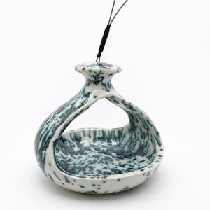 May include: A ceramic bird feeder with a unique, organic shape. The feeder is white with a speckled, teal-colored pattern. It has a loop-like design with a small opening at the top for hanging. A black cord is attached for hanging.