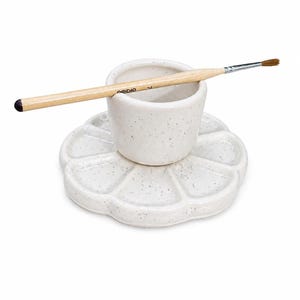 May include: A speckled white ceramic paint palette with a flower-shaped design, holding a small cup and a paintbrush. The paintbrush has a wooden handle and a fine, brown bristle head. The word "ORIDIO" is printed on the handle.