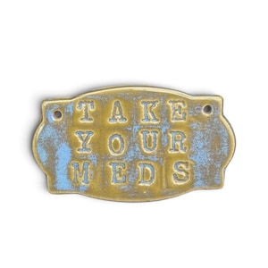 Puede incluir: Una placa metálica rectangular de color latón con bordes redondeados y dos pequeños orificios para montaje. La superficie presenta un acabado desgastado con parches azules y dorados, y el texto en relieve dice TAKE YOUR MEDS en una fuente clara.