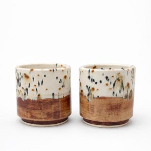 Puede incluir: Dos tazas de cer&aacute;mica con un dise&ntilde;o moteado. La mitad superior de cada taza es blanca con motas negras y marrones, mientras que la mitad inferior es marr&oacute;n liso. Las tazas son cil&iacute;ndricas con un borde ligeramente ensanchado.