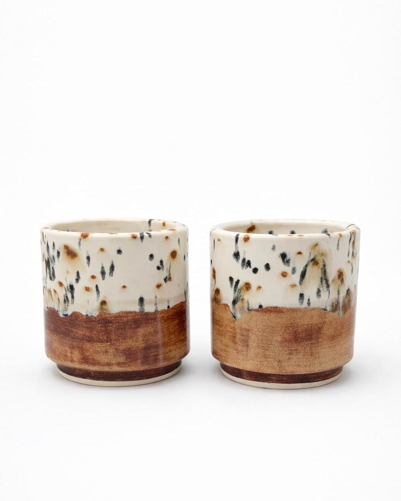 Puede incluir: Dos tazas de cer&aacute;mica con un dise&ntilde;o moteado. La mitad superior de cada taza es blanca con motas negras y marrones, mientras que la mitad inferior es marr&oacute;n liso. Las tazas son cil&iacute;ndricas con un borde ligeramente ensanchado.