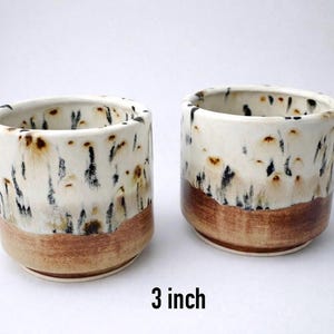 Puede incluir: Dos tazas de cer&aacute;mica hechas a mano, cada una de 7,6 cm de altura, con un esmalte blanco moteado y una base marr&oacute;n. Las tazas tienen forma cil&iacute;ndrica y una est&eacute;tica r&uacute;stica.