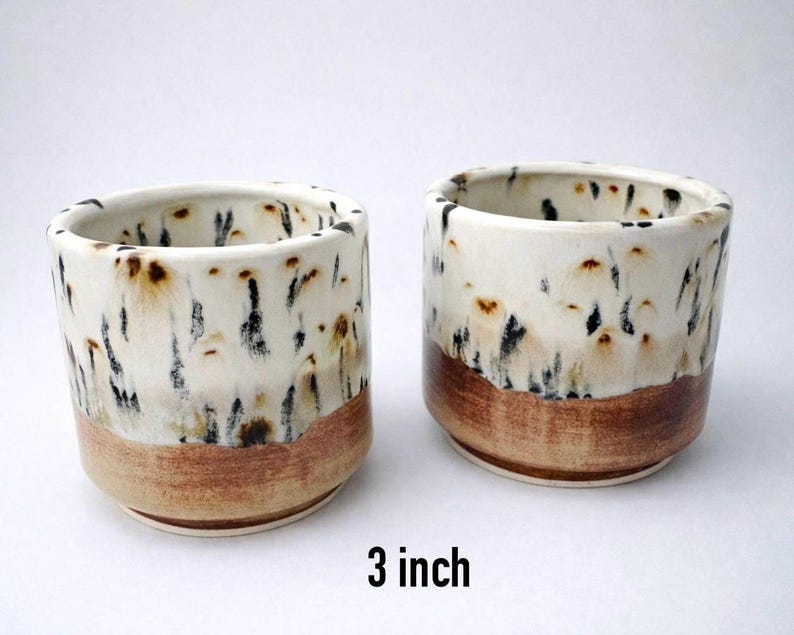 Puede incluir: Dos tazas de cer&aacute;mica hechas a mano, cada una de 7,6 cm de altura, con un esmalte blanco moteado y una base marr&oacute;n. Las tazas tienen forma cil&iacute;ndrica y una est&eacute;tica r&uacute;stica.