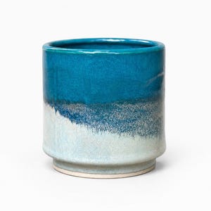 May include: A cylindrical ceramic cup featuring a glossy deep teal glaze on the upper portion that transitions into a textured, speckled light blue and white finish toward the base, which has a slightly recessed foot and a smooth, flat rim.