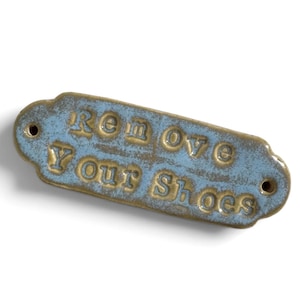 May include: A rectangular metal sign with rounded edges and two mounting holes, featuring a distressed blue finish over a bronze-colored base. The embossed text reads "Remove Your Shoes" in a stylized font, with the bronze metal showing through.