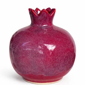 May include: A pomegranate-shaped ceramic vase with a glossy, deep magenta glaze. The vessel features a rounded body and a crown-like top opening with pointed edges. The surface displays subtle variations in color intensity and a smooth finish.