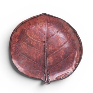 May include: A ceramic dish shaped like a leaf with a textured surface. The dish features a deep reddish-brown glaze with darker mottled accents and a prominent central vein running from the stem base to the rounded tip of the leaf-shaped form.