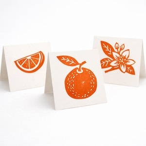 May include: Three folded greeting cards with orange illustrations. One card features a slice of orange, another an orange with a leaf, and the third a floral design. The illustrations are printed in orange on a white background.
