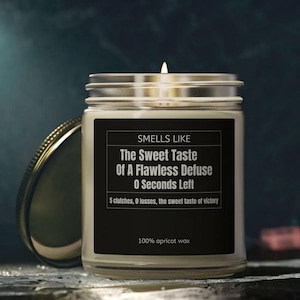 May include: A clear glass jar candle with a black label featuring white text that reads: SMELLS LIKE The Sweet Taste Of A Flawless Defuse 0 Seconds Left 5 clutches, 0 losses, the sweet taste of victory 100% apricot wax. The candle has a lit wick.
