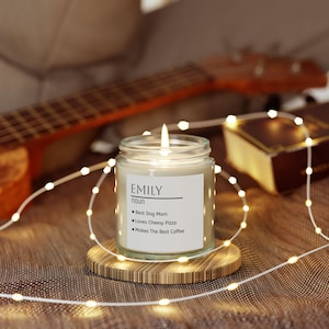 Puede incluir: Una vela encendida en un frasco de vidrio transparente con una etiqueta blanca que muestra el texto EMILY noun, Best Dog Mom, Loves Cheesy Pizza y Makes The Best Coffee. La vela descansa sobre un posavasos de madera rodeado de luces pequeñas.