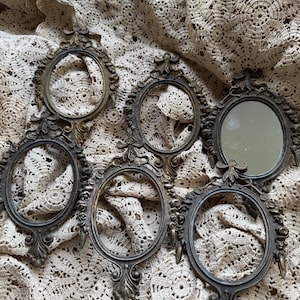 May include: An assortment of antique oval-shaped metal frames with ornate detailing. One frame contains a mirror. The frames are bronze-colored and rest on a cream-colored lace surface. The frames have decorative flourishes and a vintage aesthetic.