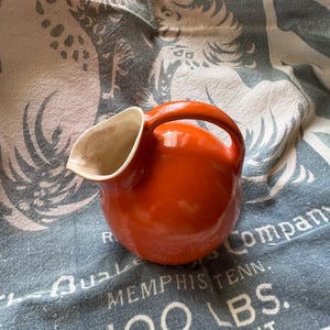 May include: A small, round, orange ceramic pitcher with a handle. The pitcher has a white interior and a spout. The pitcher is sitting on a blue and white patterned cloth. The cloth has text that reads "MEMPHIS TENN. 100 LBS. NET WEIGHT."