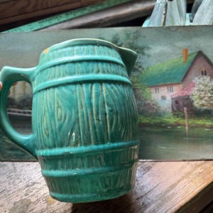 May include: A turquoise ceramic pitcher with a barrel design. The pitcher has a handle and a spout. The wood-like texture is visible on the exterior. The background includes a painting of a house and a wooden surface.