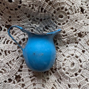 May include: A small, vintage, light blue enamel pitcher with a curved handle and a slightly flared rim. The pitcher is set against a beige, crocheted doily with a floral pattern. The pitcher has a glossy finish.