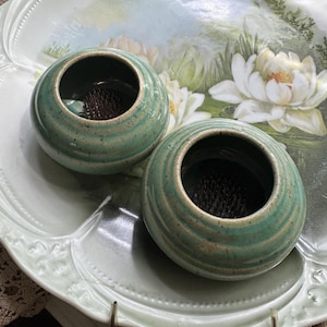 May include: Two small, round, turquoise ceramic flower frogs with a textured, ridged design. The frogs sit on a large, light green, decorative plate with a floral painting. The plate has a scalloped edge and a delicate, vintage aesthetic.